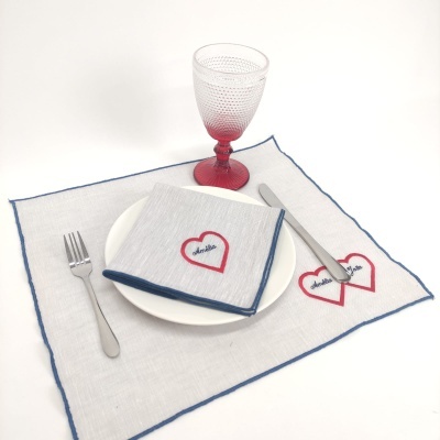 Guardanapo e tapete de mesa branco com bordas azuis e corações vermelhos bordados, prato branco, talheres e taça de vidro com haste vermelha.