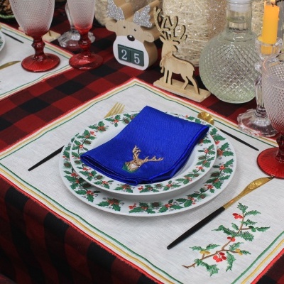 Mesa posta para jantar com decoração natalícia, incluindo pratos com folhas de azevinho, guardanapo azul e castiçais com velas amarelas.