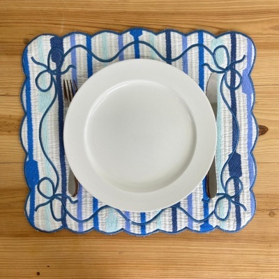 Individual de mesa têxtil azul e branco com prato branco e talheres sobre mesa de madeira