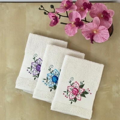 Três toalhas de mão brancas bordadas com flores coloridas ao lado de flores artificiais cor-de-rosa