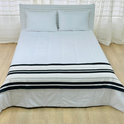 Cama com colcha branca e faixa riscada preta e cinzenta, com almofadas e cortinas brancas ao fundo