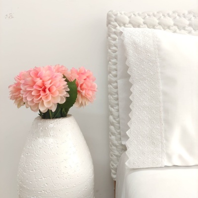 vaso branco com flores cor de rosa ao lado de cama branca com cabeceira acolchoada e almofada com bordado