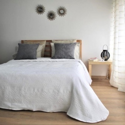 Quarto com cama com roupa de cama branca, almofadas cinzentas e mesa de cabeceira com candeeiro preto