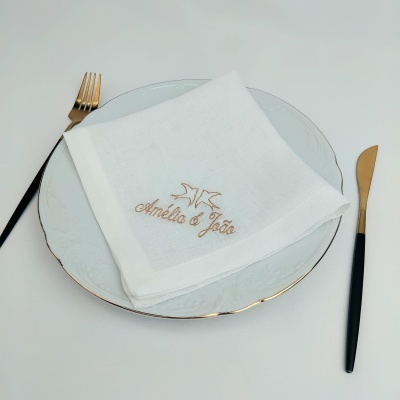 Guardanapo branco bordado com nomes sobre prato branco com bordo dourado e talheres preto e dourado
