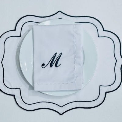 Mesa posta com jogo americano branco com bordas pretas, prato branco e guardanapo branco com letra M preta.