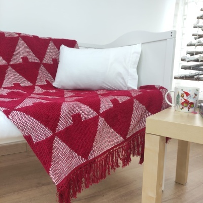Manta vermelha com padrão de triângulos brancos e franjas sobre sofá branco, almofada branca e caneca de Natal numa mesa de madeira.