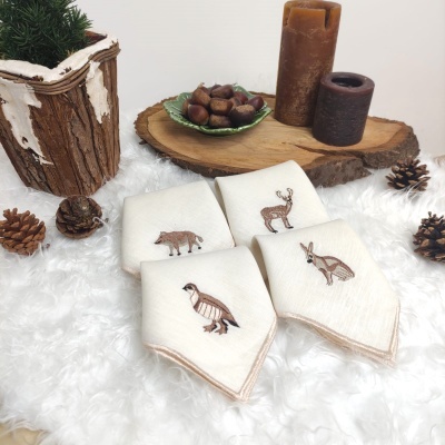 Paninhos de cozinha brancos bordados com animais sobre tecido felpudo branco, decoração rústica com velas, madeira, pinhas e castanhas.