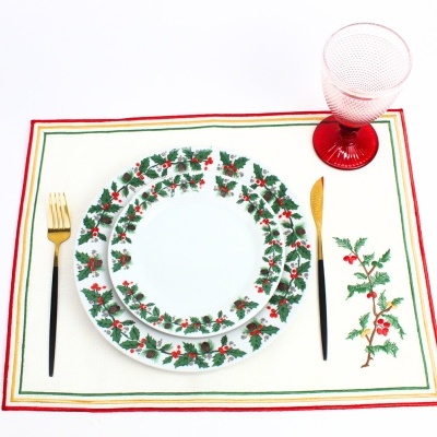 Conjunto de pratos decorados com azevinho, talheres dourados e copo vermelho sobre individual branco com bordas coloridas e desenho de azevinho