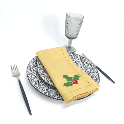 mesa posta com pratos decorados, guardanapo amarelo com azevinho, talheres pretos e copo de vidro lapidado