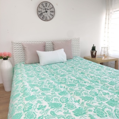 Cama com roupa de cama floral verde água e branca, almofadas cinza-bege e branca, relógio de parede, vaso com flores e mesa de madeira