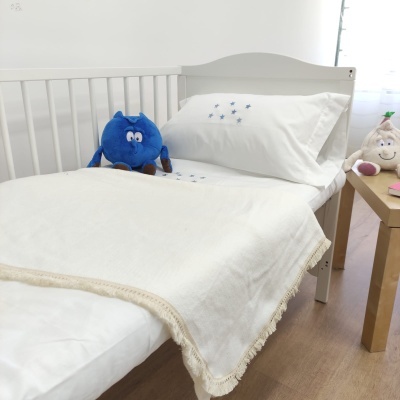 Cama de bebé branca com roupa de cama branca e manta com franjas, peluches azuis e creme próximos