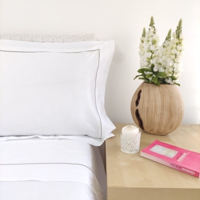 Cama com lençóis brancos e mesa de cabeceira com vaso de madeira, flores brancas, vela e livro rosa