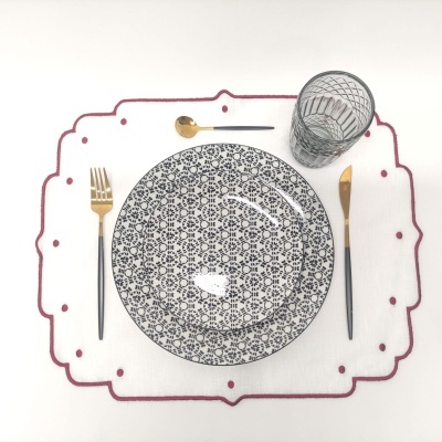 Conjunto de jantar com prato decorado, talheres dourados e copo texturizado sobre pano branco com bordado vermelho.