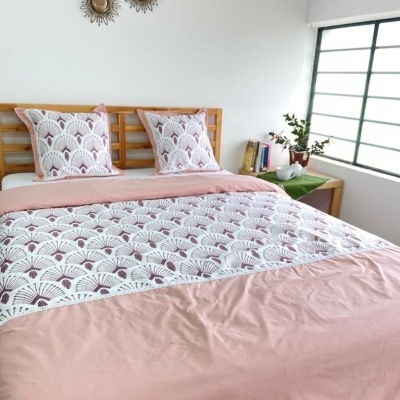 Cama com roupa de cama rosa clara e padrão branco com desenhos de leque, almofadas, mesa de cabeceira verde com livros e planta, espelhos decorativos na parede branca