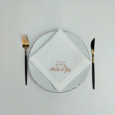 Guardanapo branco bordado com nomes Amélia e João em prato branco, garfo e faca dourados
