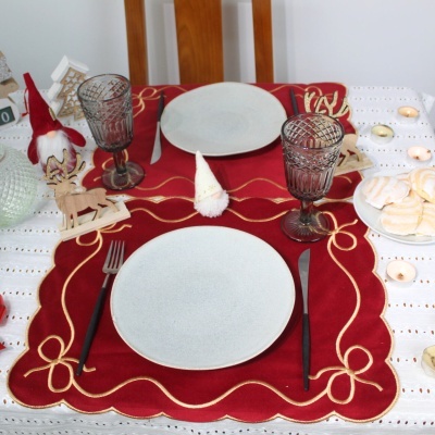 Mesa posta com toalha branca e sousplat vermelho bordado dourado, pratos brancos, copos castanhos, talheres pretos e decoração natal.