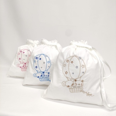 Três sacos de tecido branco bordados com balões e ursinhos em rosa, azul e bege.