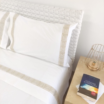 Jogo de cama branco com franjas bege geométricas mais mesa de cabeceira com candeeiro dourado e livro.