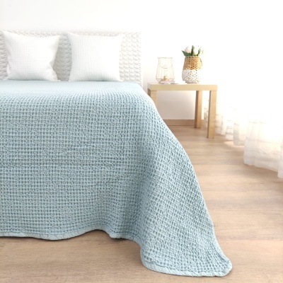 Colcha azul de malha sobre cama com almofadas brancas e mesa de cabeceira em quarto iluminado