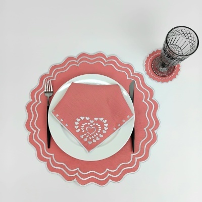 Conjunto de jantar com prato branco, guardanapo rosa com corações brancos, base de prato rosa, talheres pretos e copo com padrão de linhas em suporte rosa