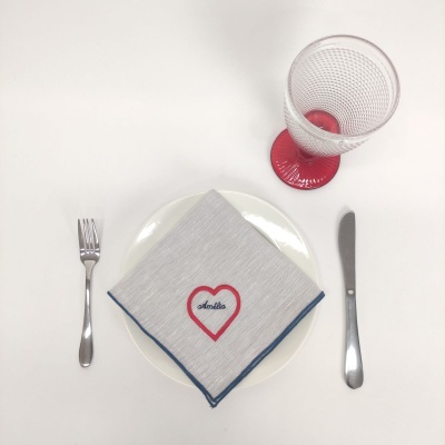 Conjunto de jantar com guardanapo cinza com coração vermelho e texto, prato branco, talheres metálicos e copo com base vermelha.