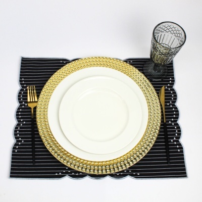 Conjunto de prato branco com prato dourado texturizado, talheres dourados e copo cinza sobre base preta com riscas brancas