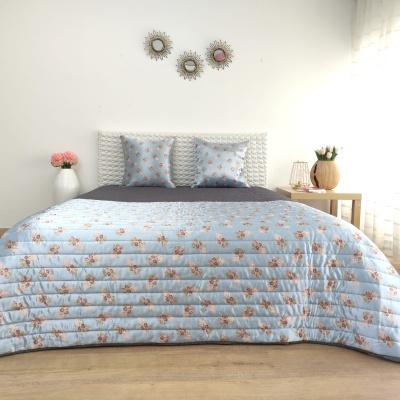 Cama com edredão azul claro com padrão floral e almofadas correspondentes, mesa de cabeceira com vaso e flores, parede branca com espelhos redondos