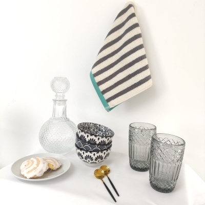 Conjunto de utensílios de mesa com garrafa, copos, tigelas, talheres, prato com doces e toalha de cozinha às riscas.