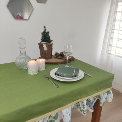 Mesa posta com toalha verde sobre toalha floral, pratos brancos, guardanapo verde, copo de vinho, velas, jarra de vidro e vaso de madeira com planta