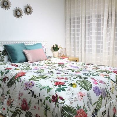 Capa de edredão floral colorida numa cama arrumada com almofadas verdes e rosa, cortinas brancas e elementos decorativos redondos na parede