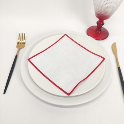 Mesa posta com pratos brancos, guardanapo com borda vermelha, talheres preto e dourado, e copo vermelho texturizado.