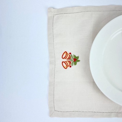 Toalhete de tecido com bordado de dois sinos vermelhos e folhas verdes, prato branco parcialmente visível