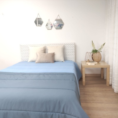 Cama com cabeceira branca trançada, edredão azul claro e almofadas bege e cinza, mesa de cabeceira madeira clara com decoração, parede branca com espelhos hexagonais.