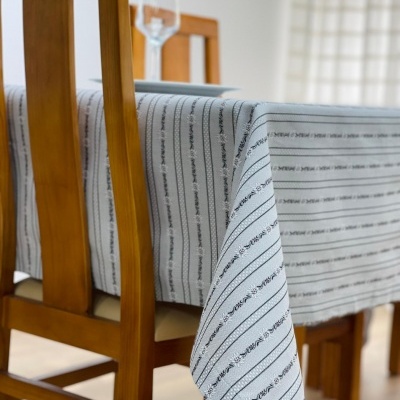 Toalha de mesa branca com riscas pretas numa mesa de madeira clara com cadeiras ao redor.