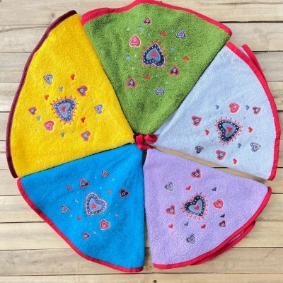Seis toalhas triangulares coloridas com borda vermelha e corações bordados, dispostas em círculo numa madeira clara.
