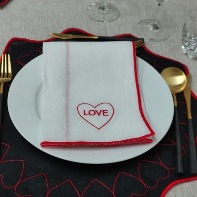 Mesa posta com jogo americano preto bordado de corações vermelhos, pratos brancos, talheres dourados e guardanapo branco com coração e palavra LOVE