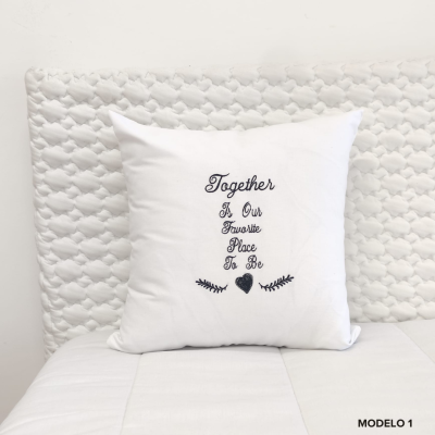Almofada branca com texto bordado a preto sobre cama branca e cabeceira estofada branca