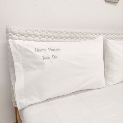 Almofadas brancas com bordado cinza numa cama com cabeceira trançada branca.