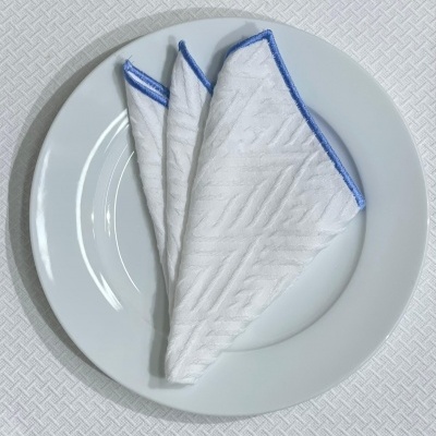 Guardanapo branco com borda azul dobrado sobre prato branco numa toalha branca texturizada