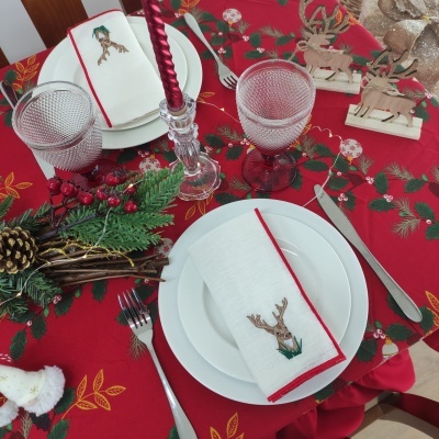 Mesa de Natal com toalha vermelha, pratos brancos, guardanapos decorados, copos texturizados, castiçal com vela, decorações de rena e arranjo de pinhas