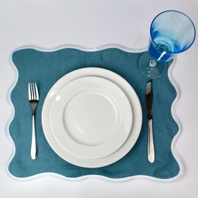 Lugar de mesa com pratos brancos, talheres prateados e copo azul sobre individual azul com borda branca ondulada