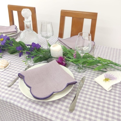 Mesa posta com toalha xadrez lilás e branco, guardanapos lilás, prato branco, talheres, copos, jarra decorativa, vela acesa e flores roxas