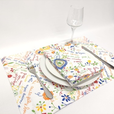 Mesa posta com toalha e guardanapo brancos com padrão floral e texto colorido, talheres e taça de vidro