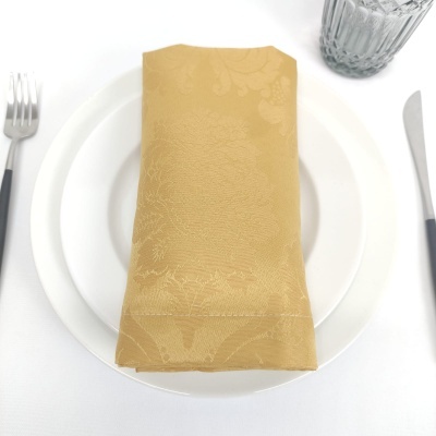 Guardanapo dourado com padrão floral sobre pratos brancos, talheres e copo em mesa branca