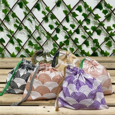 Cinco saquinhos de tecido coloridos com padrão geométrico em bancada de madeira e fundo de treliça com folhas.