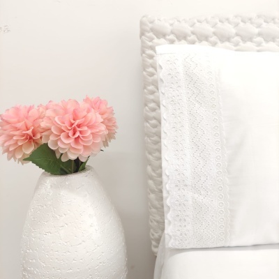 Vaso branco com flores cor-de-rosa ao lado de almofada branca com bordado rendado e cabeceira acolchoada cinzenta.