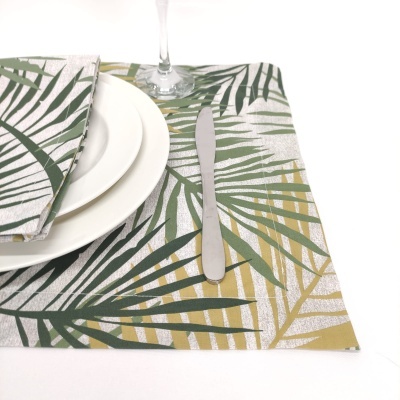 Jogo americano e guardanapo com padrão de folhas verdes e amarelas sobre mesa branca com prato branco, faca e copo