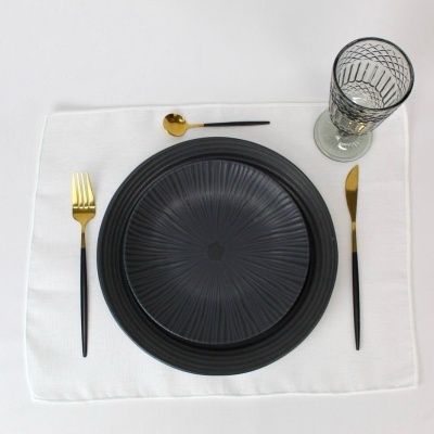 Conjunto de jantar com pratos pretos texturizados, talheres pretos e dourados, copo de vidro com padrão geométrico e toalhete branco.
