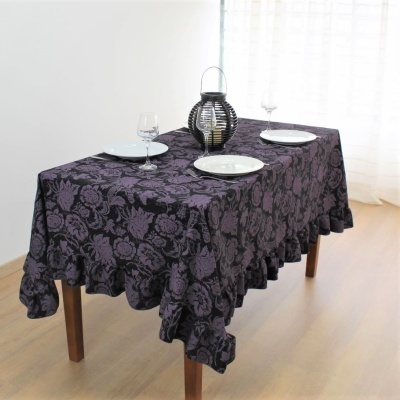 Toalha de mesa roxa com padrão floral negro sobre mesa de madeira clara, pratos brancos, copos e castiçal
