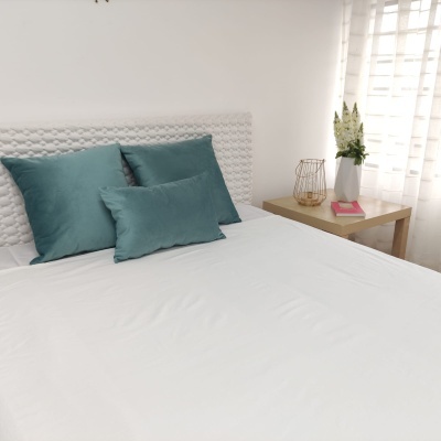 Cama com cabeceira branca e almofadas verdes, mesa de cabeceira com jarra branca, flores, candeeiro dourado e livro rosa.